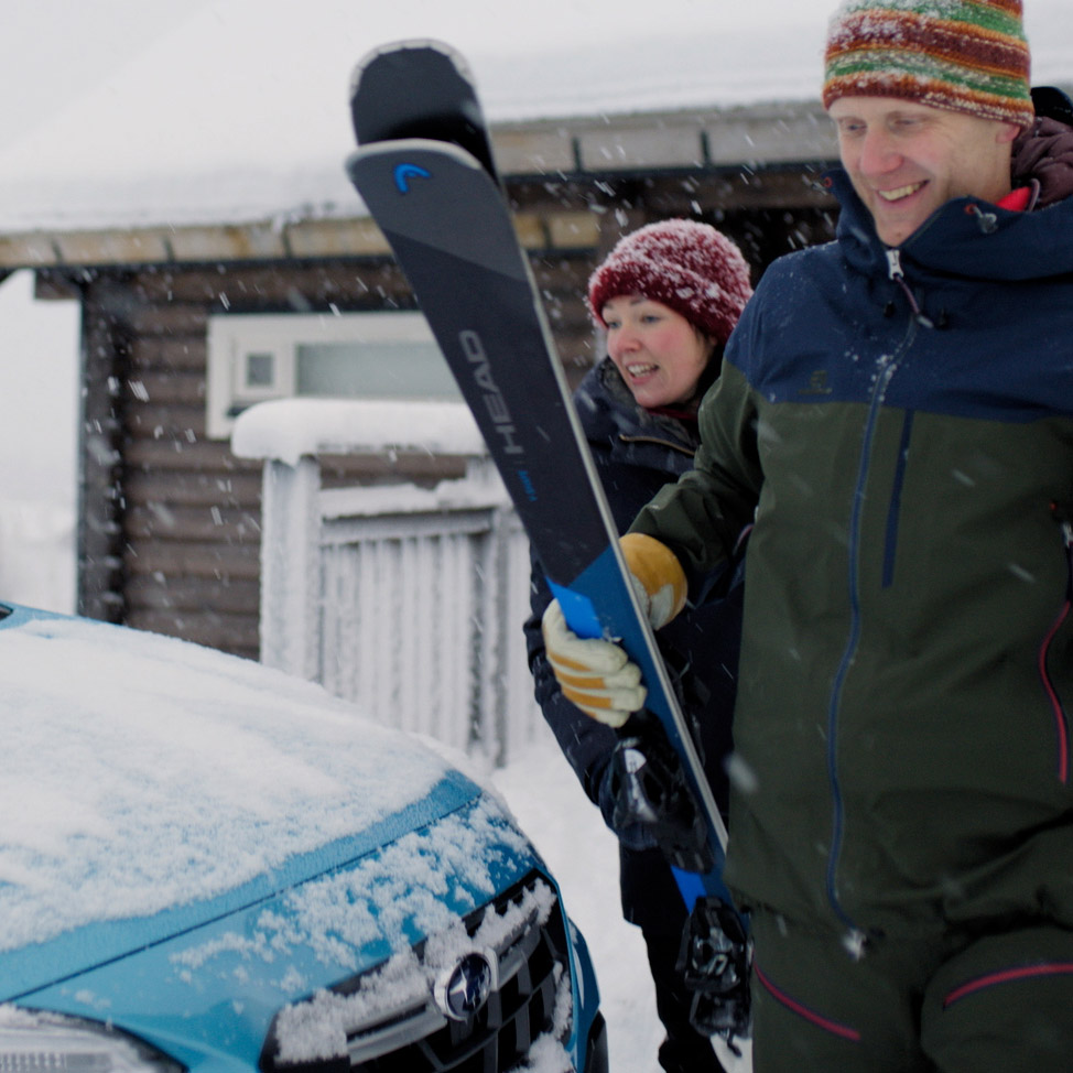 Två personer med skidor till hands bredvid Subaru i snömiljö
